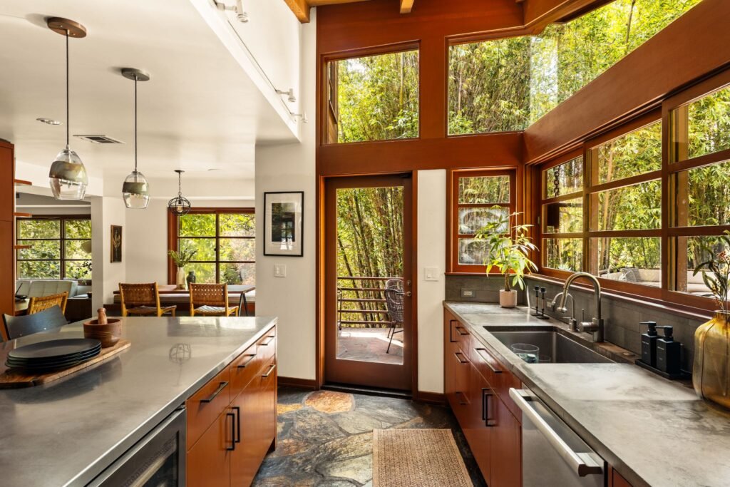 Bright mid-century modern kitchen with a large center island, slate flagstone floors, custom wood-framed glass doors, and high clerestory windows offering private bamboo forest views.