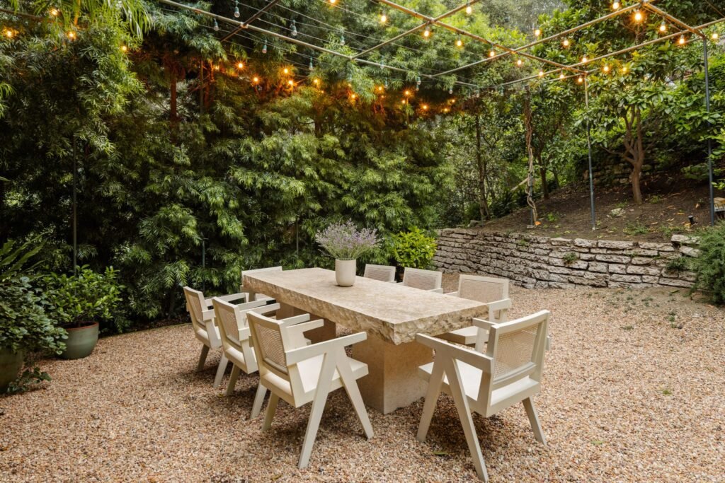 Secluded luxury outdoor dining area featuring a custom stone table, modern white Pierre Jeanneret style chairs, overhead bistro string lights, and a private gravel courtyard.