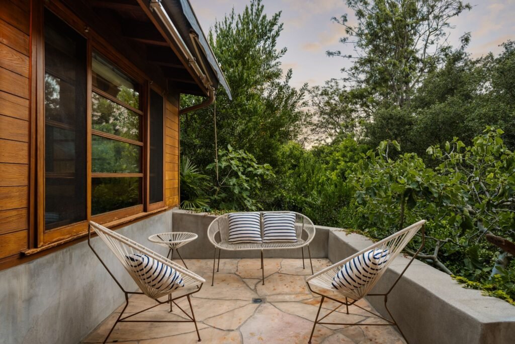 Private mid-century modern terrace featuring a flagstone floor, concrete retaining walls, white woven Acapulco-style outdoor furniture with striped pillows, and a lush canyon view.