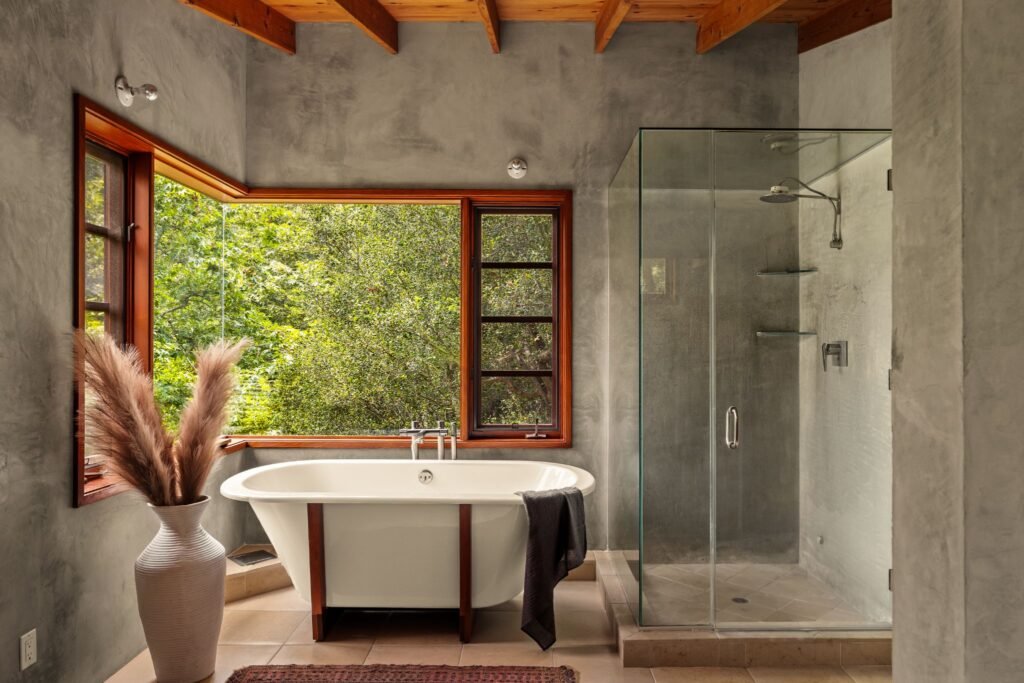 Modern luxury bathroom featuring a freestanding soaking tub next to a large corner window with lush forest views, a frameless glass shower, concrete-finish walls, and exposed wood ceiling beams.