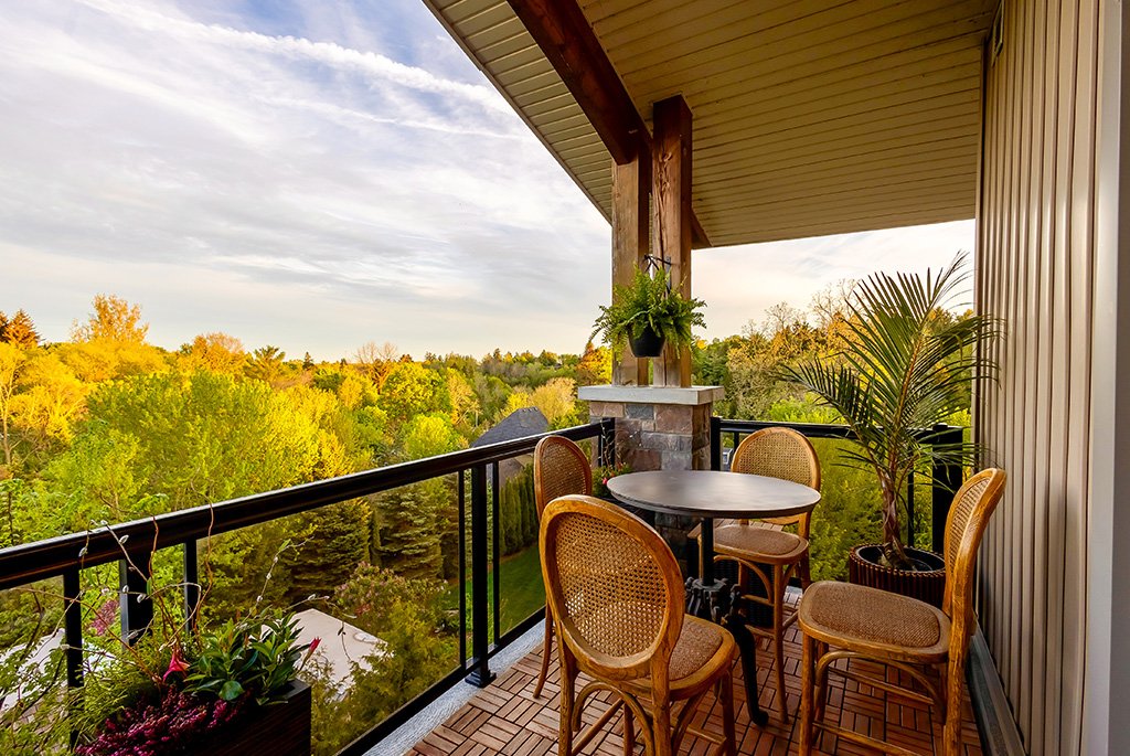 Covered condo balcony featuring interlocking wood deck tiles, a bistro dining set with woven wicker chairs, and panoramic views of lush green treetops.