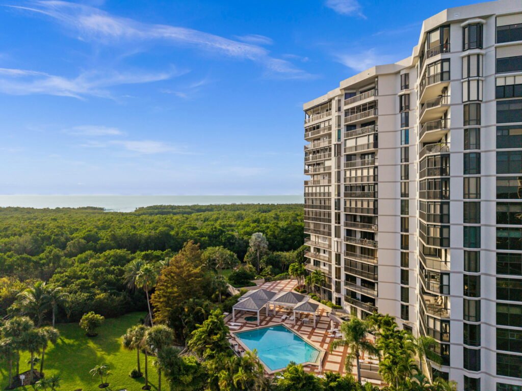 Luxury high-rise condominium building with private balconies overlooking an outdoor swimming pool, lush tropical landscaping, and a panoramic ocean view.