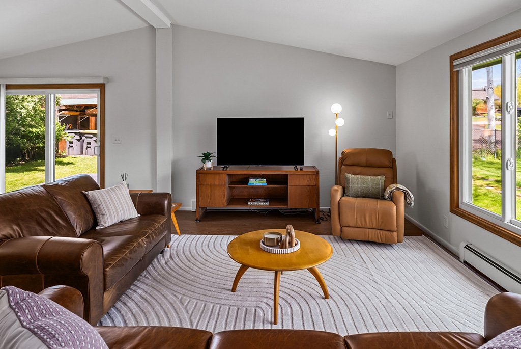 Bright living room with a vaulted ceiling, brown leather sofas, a mid-century modern wooden media console, and a large round coffee table on a textured area rug.