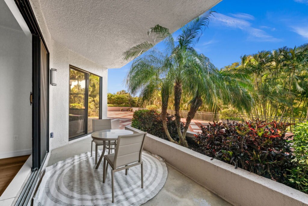 Covered condo patio balcony with a round glass table and mesh dining chairs on a round patterned outdoor rug, framed by a concrete barrier wall.
