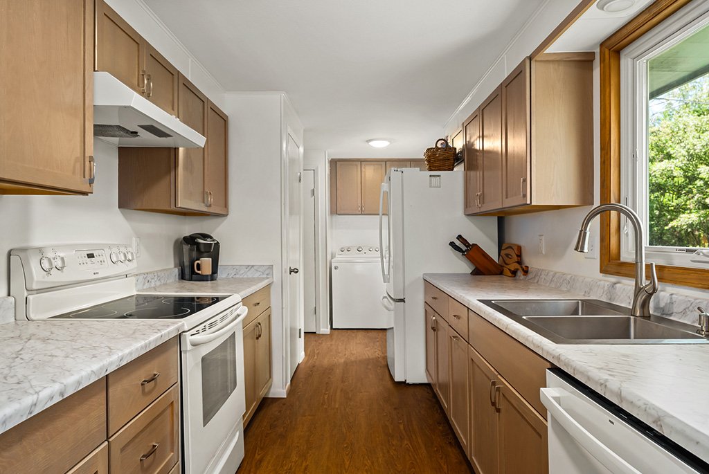 Galley kitchen layout with wood cabinets, laminate marble countertops, white appliances, a double-basin sink, and wood-look laminate flooring.