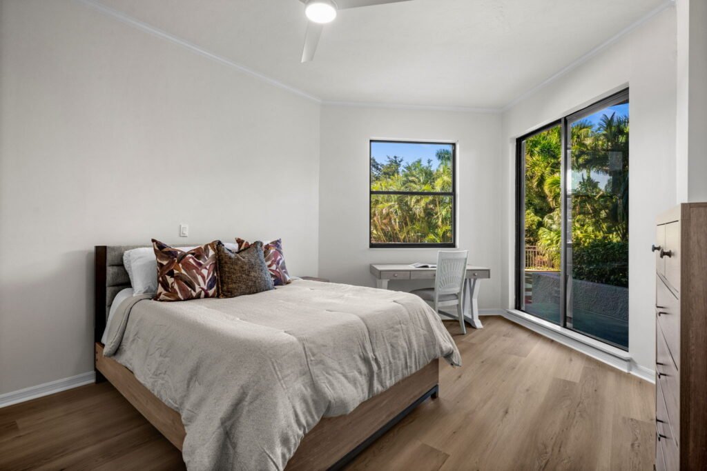 Modern bedroom featuring a wooden bed frame, light wood-plank flooring, a writing desk workspace, and large glass sliding doors opening to a tropical patio view.