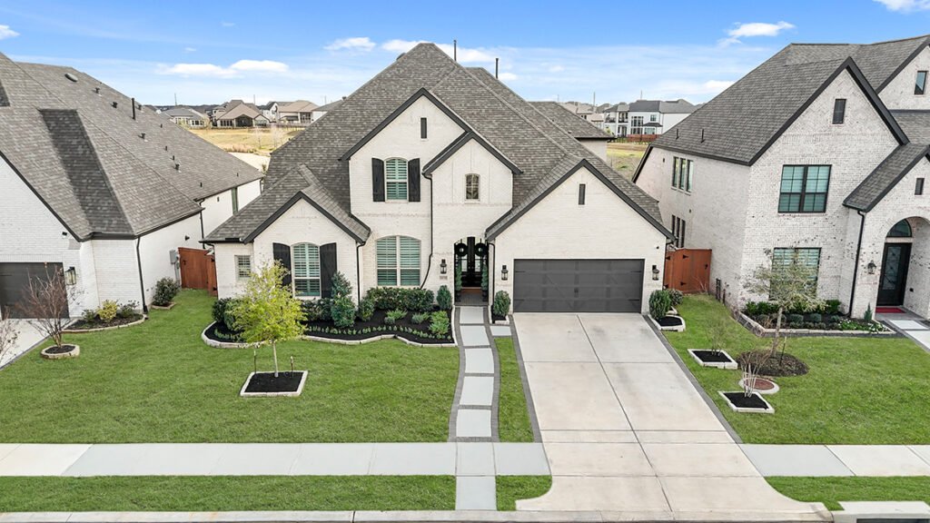 Two-story white brick traditional home with a dark grey shingle roof, two-car garage, concrete driveway, and custom stone walkway across a manicured lawn.
