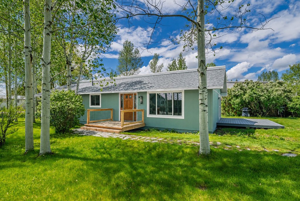 Single-story green ranch home with a wood-plank front deck, hog wire railings, asphalt shingle roof, and a stone walkway.