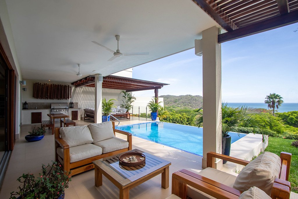 Covered patio deck with teak wood lounge chairs, an outdoor kitchen barbecue station, and a private infinity swimming pool overlooking the ocean.