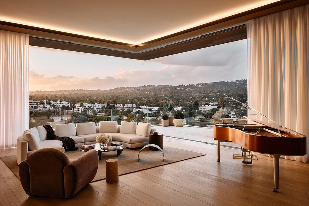 A luxury penthouse living room at 8899 Beverly Boulevard in West Hollywood featuring an open floor plan, a custom grand piano, a large beige sectional sofa, and floor-to-ceiling sliding glass walls that open to a panoramic view of the Los Angeles landscape.