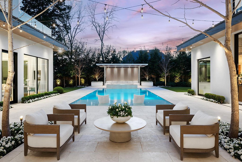 A luxury resort-style backyard patio designed by DDLA Design at a private residence in Highland Park, Dallas, featuring an integrated swimming pool, an open pavilion, wood-framed lounge chairs, and overhead string lights at dusk.