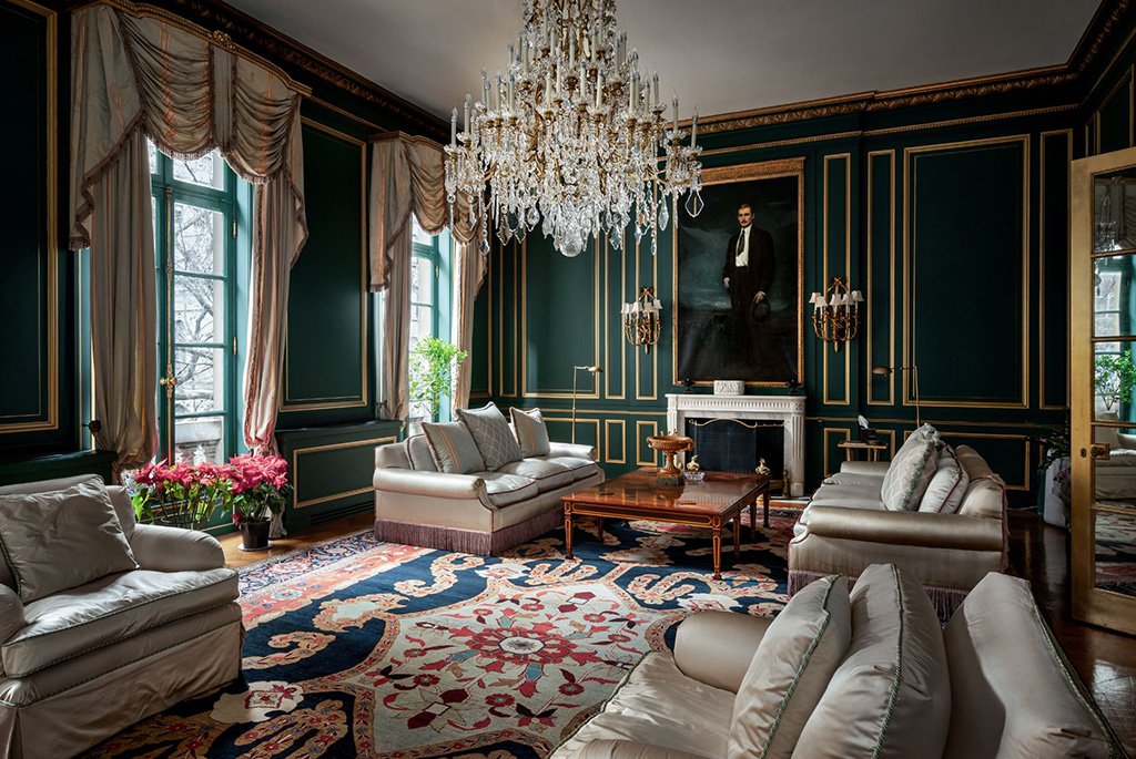 The formal drawing room of the historic Beaux-Arts style James F. D. Lanier House in New York City, featuring dark green walls with gold molding, a crystal chandelier, elegant silk sofas, and a large patterned area rug.