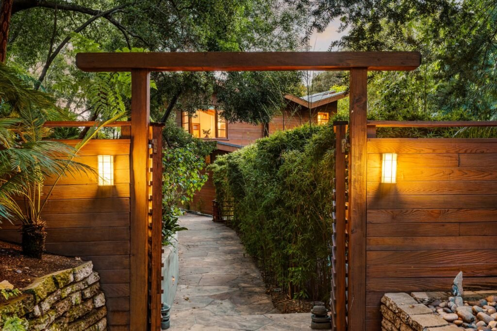 Luxury real estate entrance featuring a custom wood gate with warm sconce lighting and a stone pathway leading to a secluded mid-century modern home.