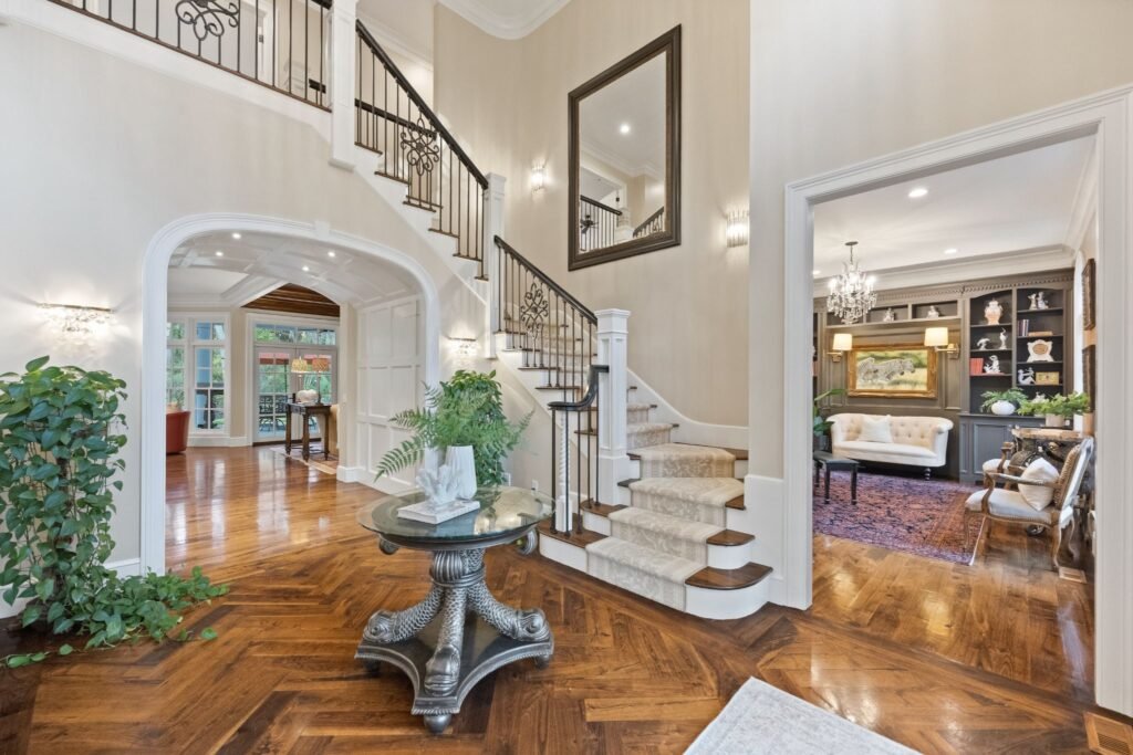 A grand home foyer featuring a curved staircase, patterned hardwood floors, a round center table, and a view into an adjacent sitting room.