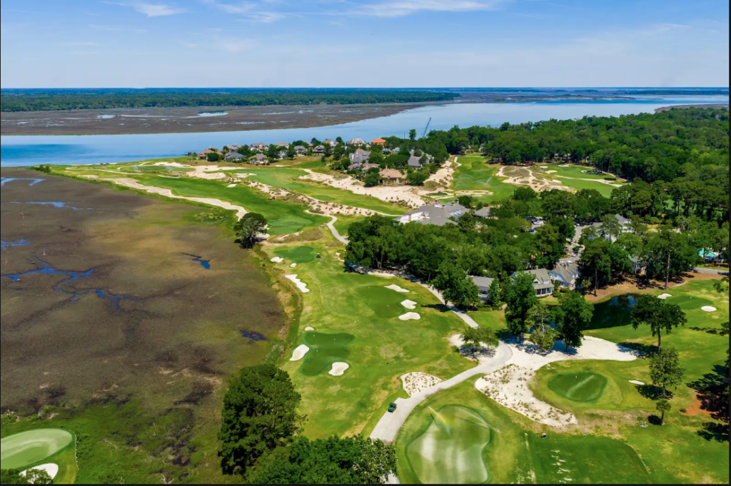 Aerial view of a coastal golf course community featuring manicured greens and fairways alongside a wide river and tidal marshlands.