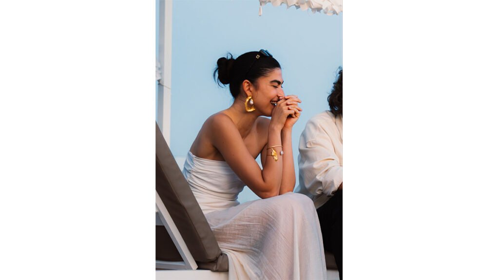 A smiling young woman in a white strapless maxi dress sits outdoors, wearing prominent gold statement earrings, a matching gold cuff bracelet, and her hair up in a sleek bun