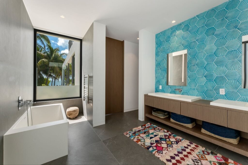 Luxurious primary bathroom at 19 Avacado Street in Islamorada, featuring a modern standalone tub, a large picture window with natural light, and a double floating vanity against a blue geometric tile accent wall.
