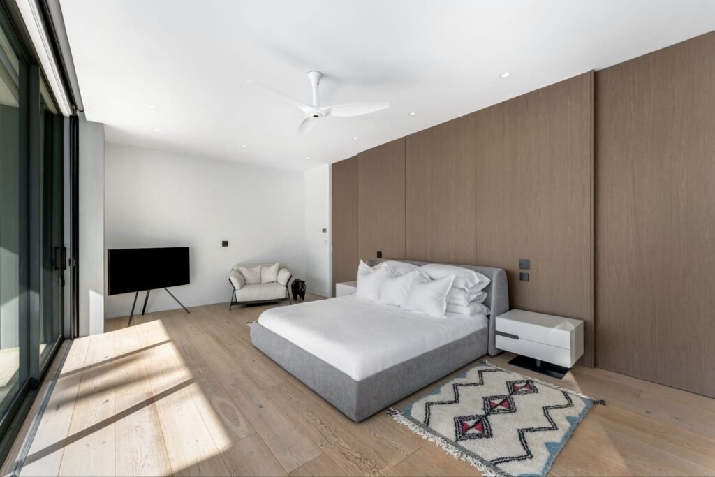 First floor primary suite at 19 Avacado Street in Islamorada, featuring a modern grey bed, engineered hardwood flooring, a minimalist wood accent wall, and retractable glass patio doors.