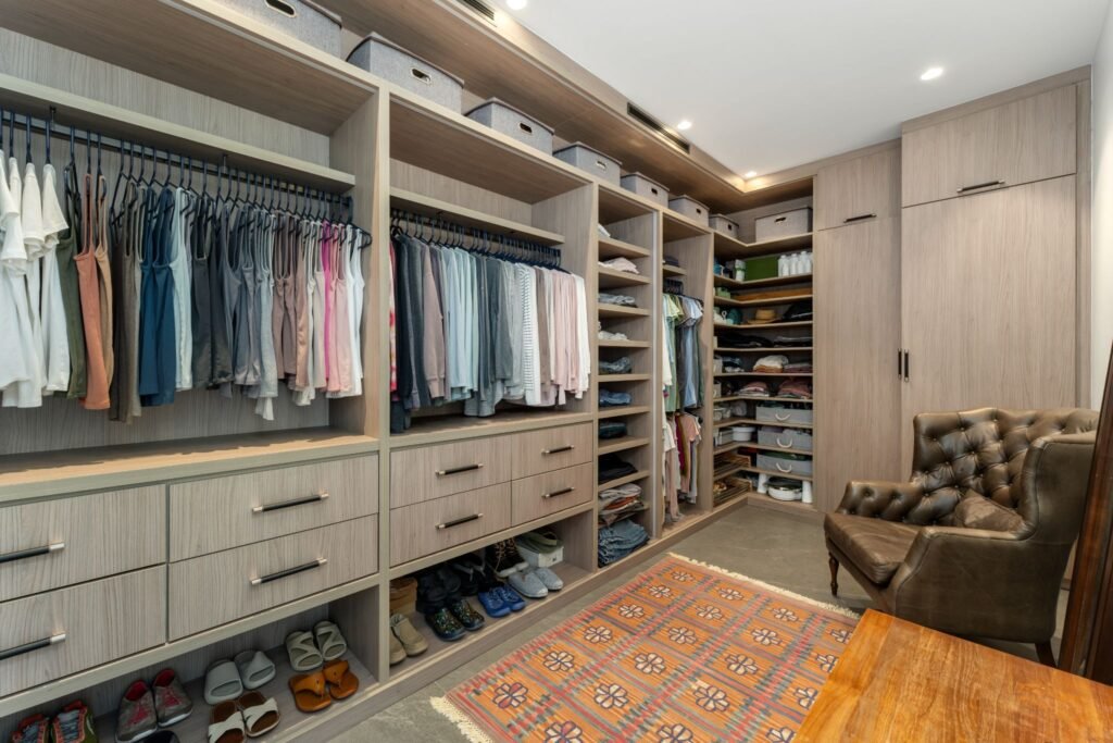 Large primary walk-in closet at 19 Avacado Street in Islamorada, showcasing custom built-in wood shelving, organized clothing racks, shoe storage, and a leather accent armchair.