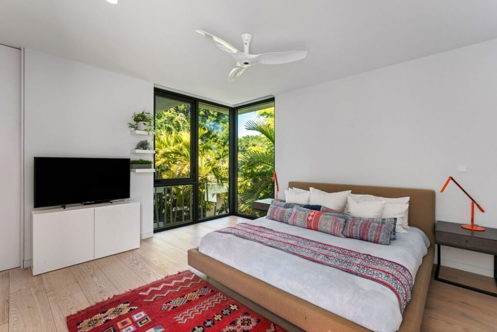 Private upstairs ensuite bedroom at 19 Avacado Street in Islamorada, featuring engineered hardwood flooring, a modern fabric bed frame with patterned bedding, and floor-to-ceiling corner windows framing tropical palm views.