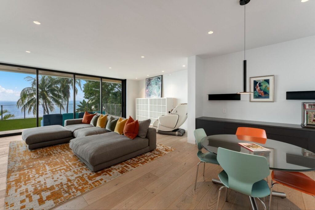 Upstairs open floor plan living area at 19 Avacado Street in Islamorada, featuring an L-shaped grey sectional sofa, engineered hardwood floors, a round dining table, and glass doors opening to elevated bay views.