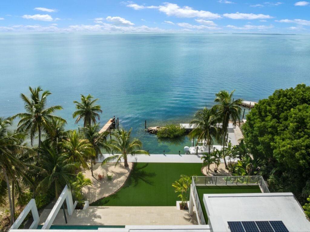 Aerial drone view of the waterfront estate at 19 Avacado Street in Islamorada, showing panoramic sunset-facing bay views, a private boat dock, a green artificial turf lawn, and tropical palms.