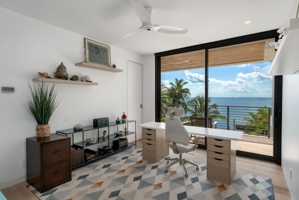 Modern home office bedroom at 19 Avacado Street in Islamorada, featuring a light wood desk, contemporary desk chair, geometric patterned rug, and floor-to-ceiling glass doors opening to panoramic bay views.