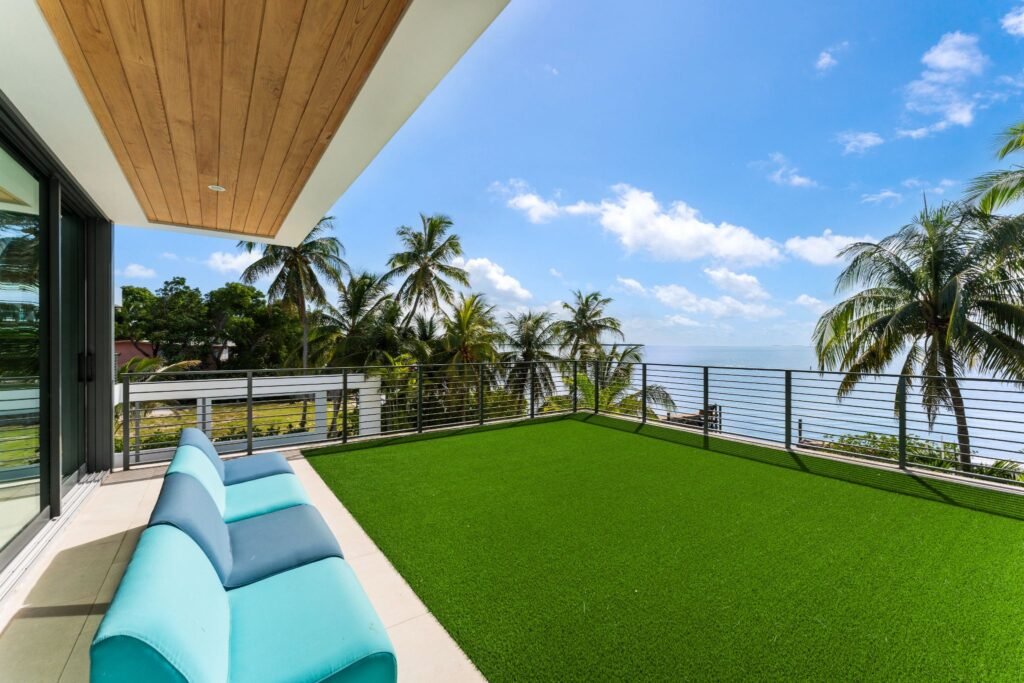Elevated rooftop terrace at 19 Avacado Street in Islamorada, featuring green artificial turf, a blue contemporary outdoor sectional sofa, and panoramic bay views framed by tropical palms.