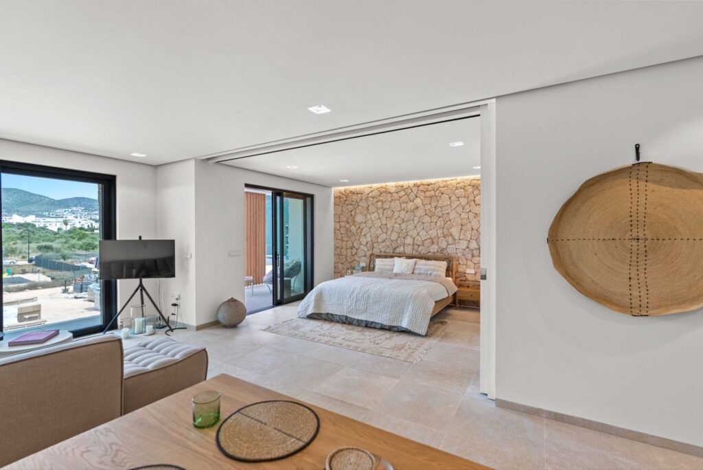 Modern 1-bedroom apartment bedroom in Playa d’en Bossa, Ibiza, featuring sliding pocket doors, a textured stone accent wall, and an open-plan layout.