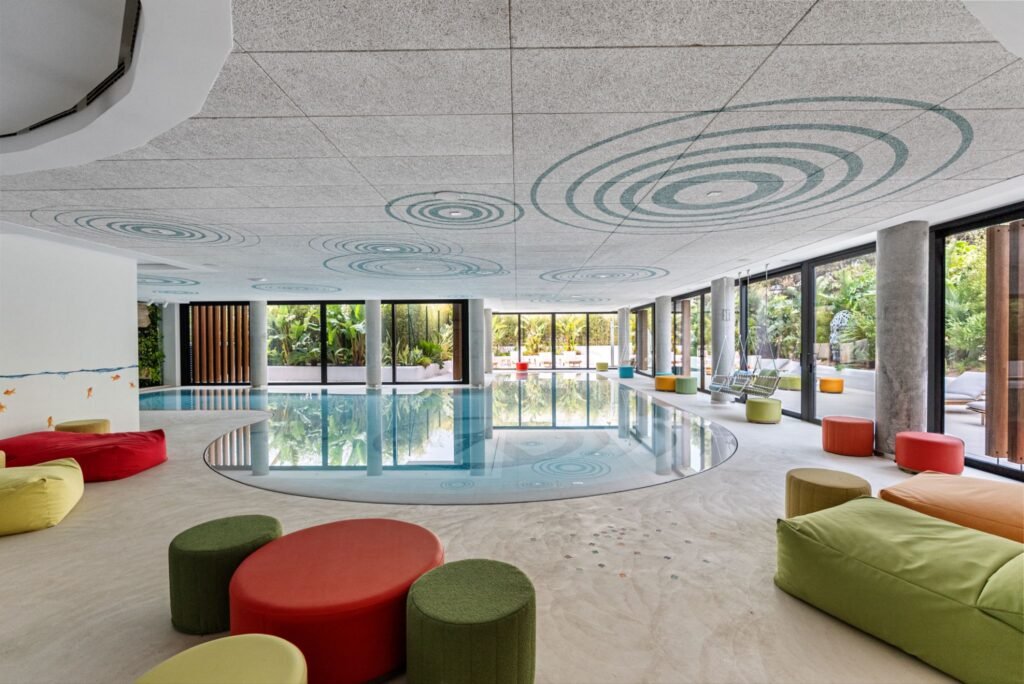 Luxury apartment building indoor pool and lounge area in Playa d’en Bossa, Ibiza, with colorful seating, floor-to-ceiling windows, and tropical outdoor views.