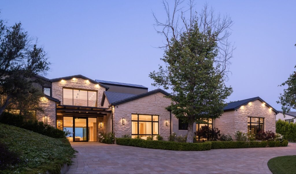 Luxury modern estate exterior at dusk featuring an illuminated stone facade, custom glass entryway, and a sprawling paved driveway.