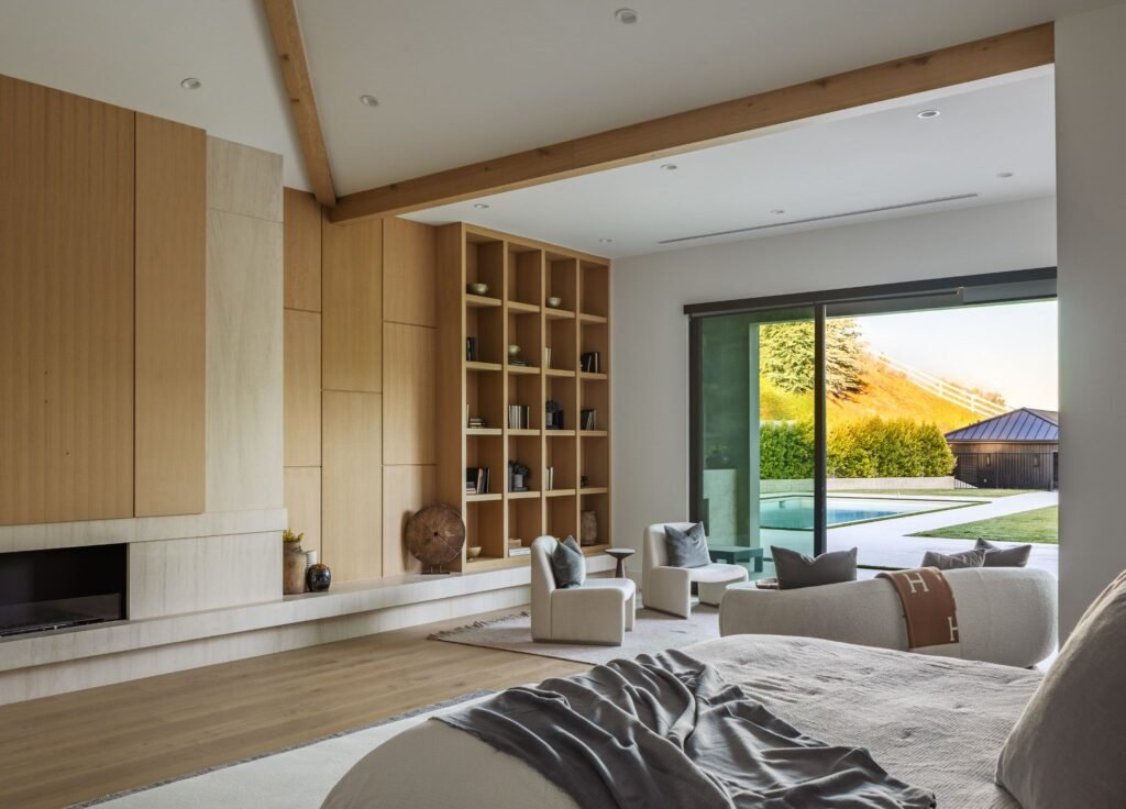 Modern luxury master bedroom suite with a sitting area, floor-to-ceiling wood built-in shelving, and large sliding glass doors leading to a backyard pool.