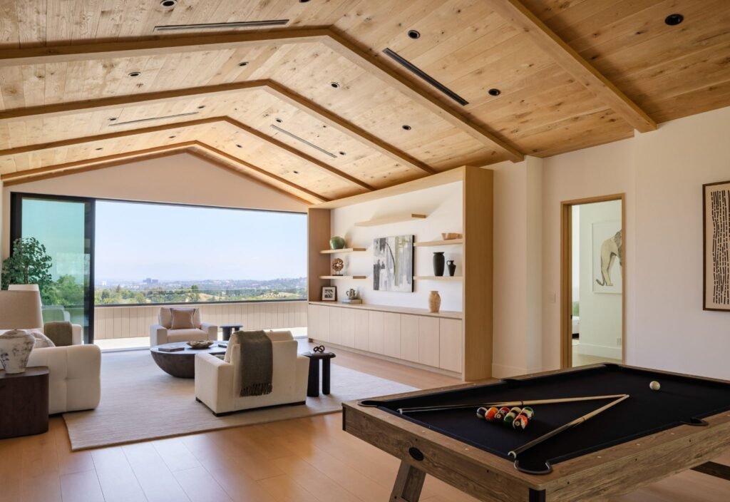 Luxury game room featuring a black-felt pool table, modern sitting area, and built-in shelving under a vaulted wood ceiling with panoramic views.