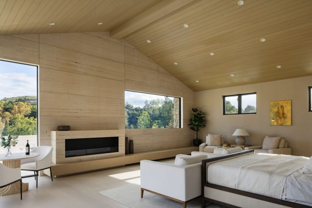 Luxury master bedroom featuring a modern linear fireplace set into a travertine stone wall under a vaulted wood-paneled ceiling.