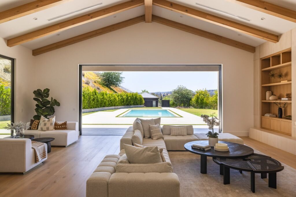Luxury open-concept living room with a large pocket glass door opening to a backyard swimming pool under a vaulted beamed ceiling.