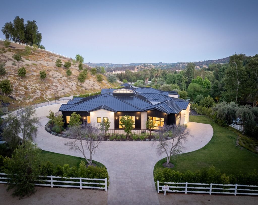 Aerial view of a luxury modern farmhouse estate featuring a black standing-seam metal roof, circular paved driveway, and rolling green hills.