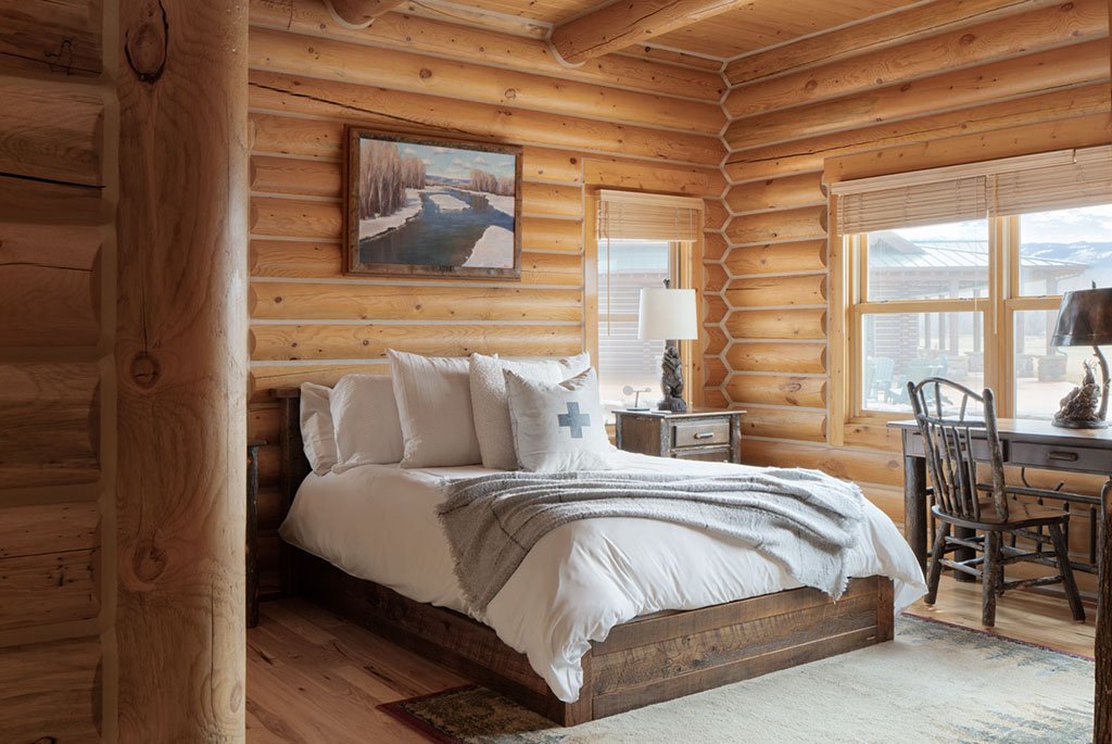 A guest bedroom at A River's Ranch featuring golden hand-hewn logs, a rustic wooden desk, and large windows looking out toward the estate grounds.