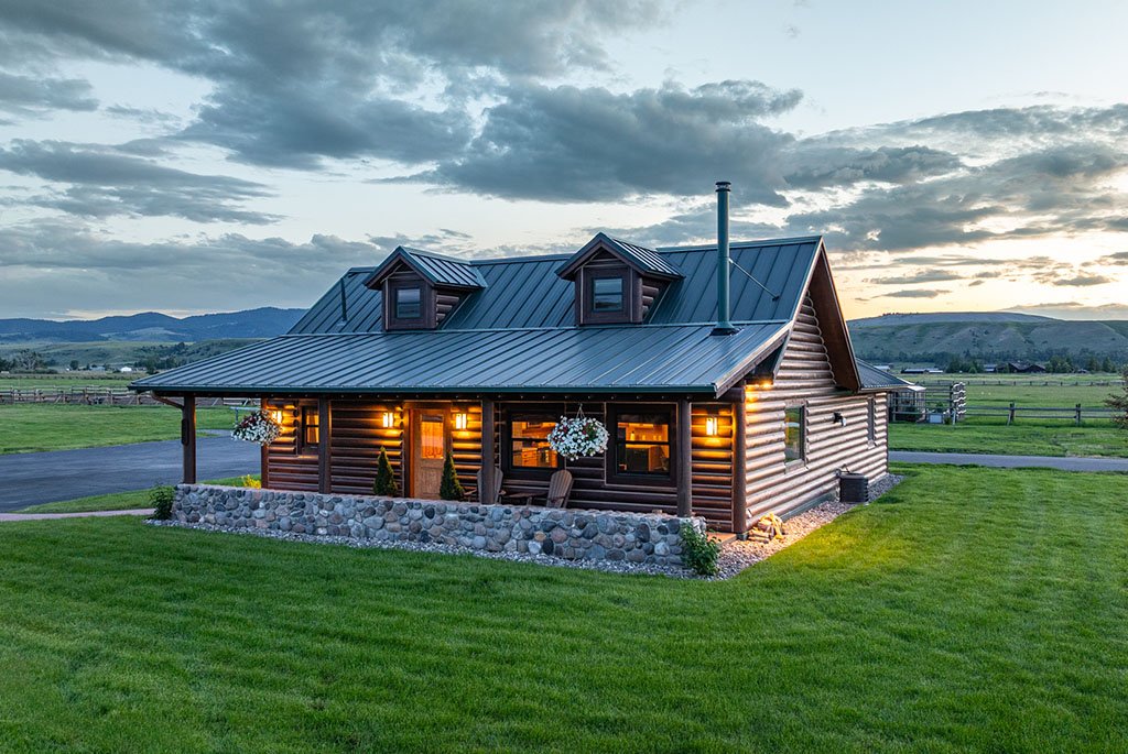 The private one-bedroom guest house at A River's Ranch featuring a dark green metal roof, a stone-lined patio, and a log exterior surrounded by pastures.