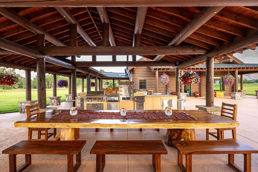 The covered outdoor kitchen at A River's Ranch featuring a long live-edge wood dining table, built-in smoker, and a premium grill setup for al fresco entertaining.