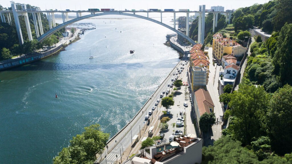 Aerial riverfront view of the Prisma Douro residential development area along the Douro River in Porto's Foz do Douro, featuring the iconic Arrábida Bridge and waterfront roads.