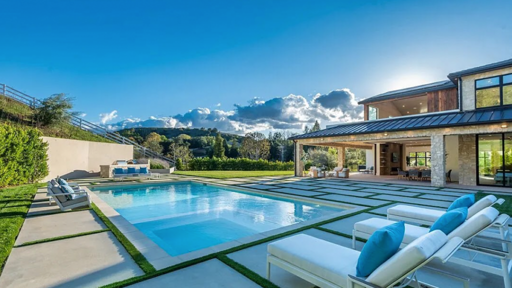 Luxury backyard patio with a rectangular swimming pool, modern white lounge chairs with blue pillows, and large grid pavers.