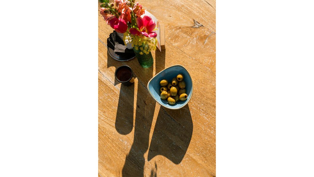 Top-down view of a sunny wooden table with a blue ceramic bowl of green olives, a small cup, and bright pink and yellow flowers in a green glass vase casting long shadows.