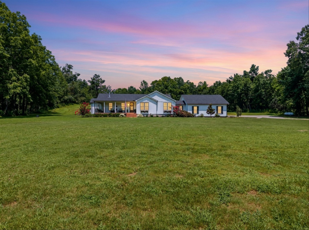 A front view of the custom modern farmhouse residence at 121 Harbor Lane in Hickory Creek, Texas, situated on an organic multi-generational family compound with a large green lawn, surrounded by dense trees under a sunset sky.