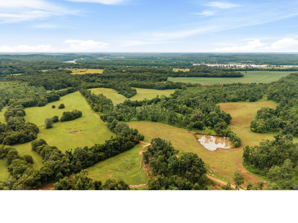An aerial view of the expansive 144-acre organic farm compound at 121 Harbor Lane in Hickory Creek, Texas, showing rolling green cattle pastures, a private pond, wetlands, and mature timber acreage under a clear sky.