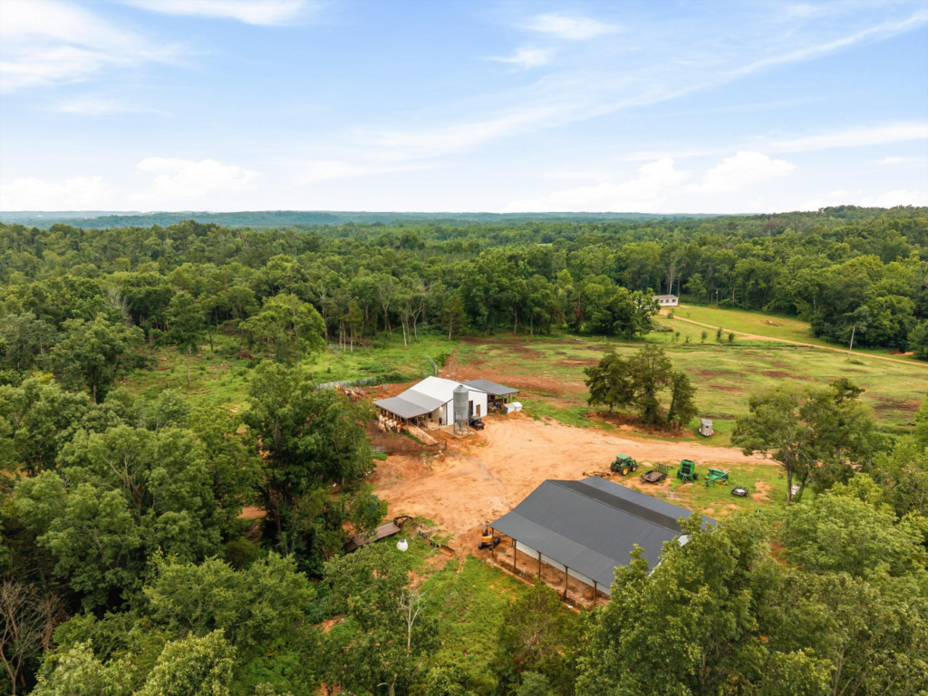 An aerial view of the active Wagyu cattle ranching operations at 121 Harbor Lane in Hickory Creek, Texas, featuring large agricultural barns, silos, ranch equipment, and open organic pastureland surrounded by dense mature timber.