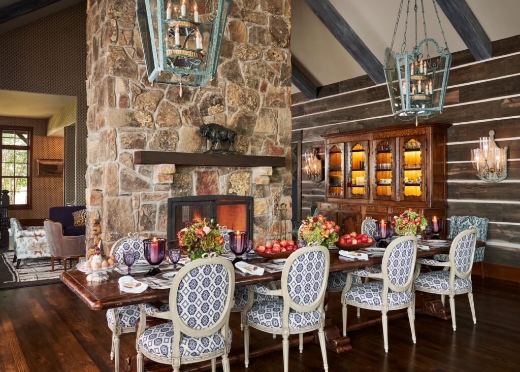 The luxury rustic dining room interior at the 50-acre legacy farmhouse estate outside Dallas, Texas, custom designed by Kirsten Kelli Interior Design, featuring a large stone fireplace, exposed log walls, a long wooden dining table with patterned chairs, and unique hanging cage chandeliers.