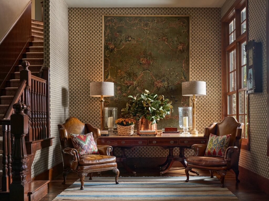 A custom-designed foyer sitting area at the 50-acre legacy estate outside Dallas, Texas, curated by Kirsten Kelli Interior Design, featuring a wood staircase, patterned wallpaper, a central console table, leather wingback chairs, and large tapestries.