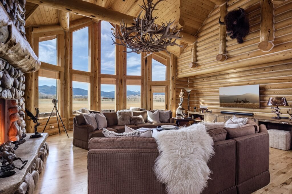 The luxury rustic great room interior of the 6,612-SF main log home at A River’s Ranch in Gallatin Gateway, Montana, featuring a massive stone fireplace, an antler chandelier, exposed log walls, a large sectional sofa, and expansive floor-to-ceiling windows showing mountain views.