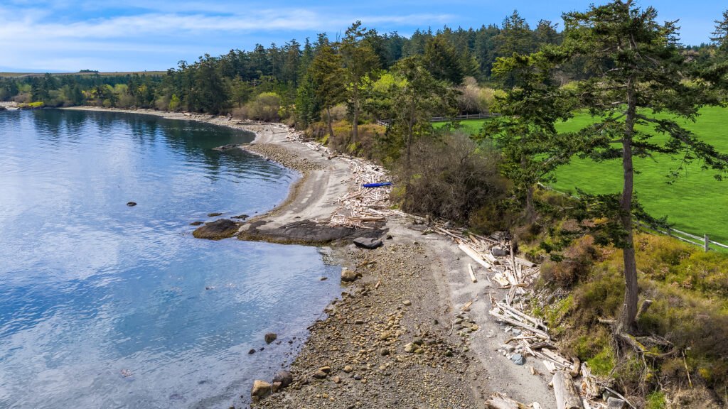 An aerial view of the untamed beach frontage at Halftide Farms on San Juan Island, Washington, showcasing a rocky shoreline, driftwood, clear blue ocean water, and adjacent green pastures bordered by a mature evergreen forest.