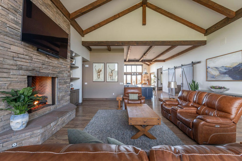 The open-concept living room inside the integrated secondary residence at 61465 White Peaks Court in Bend, Oregon, featuring high vaulted ceilings with custom wood beams, a stone fireplace, brown leather sofas, and sliding barn doors.